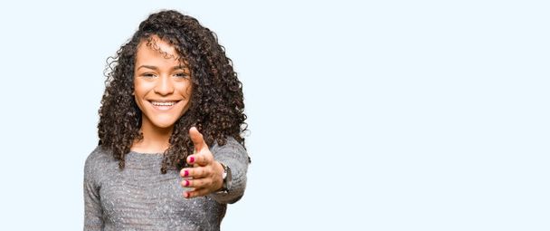 Mujer hermosa joven con el pelo rizado usando suéter gris sonriendo amistoso ofreciendo apretón de manos como saludo y bienvenida. Negocios exitosos
. - Foto, Imagen