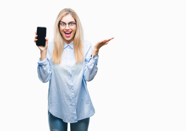 Young beautiful blonde business woman showing screen of smartphone over isolated background very happy and excited, winner expression celebrating victory screaming with big smile and raised hands - Foto, afbeelding