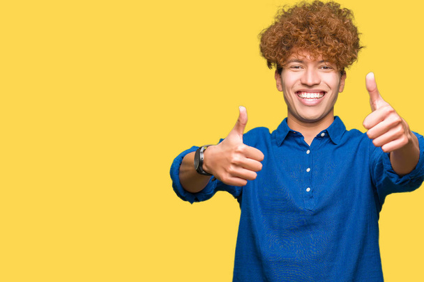 Young handsome elegant man with afro hair approving doing positive gesture with hand, thumbs up smiling and happy for success. Looking at the camera, winner gesture. - Фото, изображение