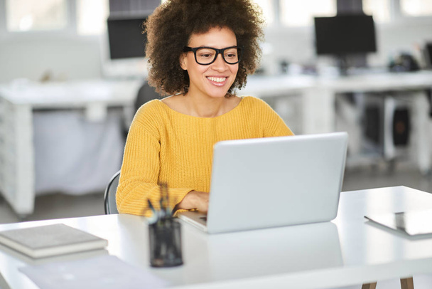 Beautiful smiling mixed race businesswoman dressed casual sitting in office and using laptop. - Photo, Image
