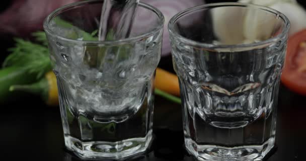 Giet alcohol wodka uit een fles in twee shot glas. Achtergrond met groenten - Video