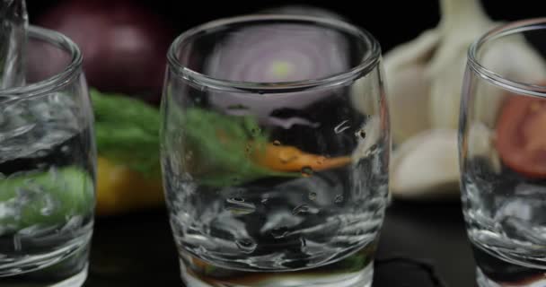 Pour alcohol vodka from a bottle in two shot glass. Background with vegetables - Footage, Video