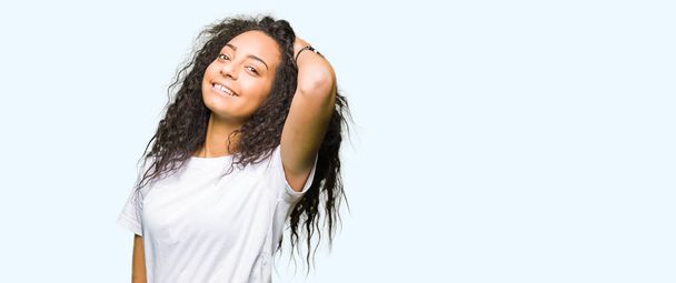 Молода красива дівчина з кучерявим волоссям в повсякденній білій футболці Усміхаючись впевнено торкається волосся ручним жестом, позує привабливо
 - Фото, зображення