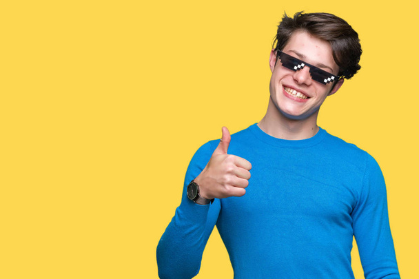 Young man wearing funny thug life glasses over isolated background doing happy thumbs up gesture with hand. Approving expression looking at the camera with showing success. - Photo, Image