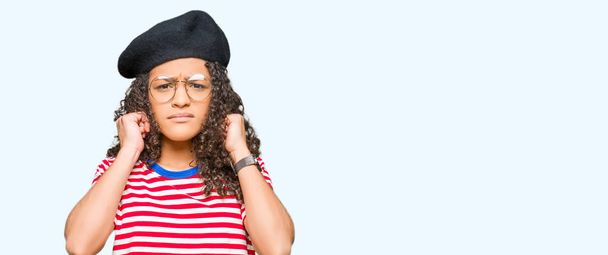 Young beautiful woman with curly hair wearing glasses and fashion beret covering ears with fingers with annoyed expression for the noise of loud music. Deaf concept. - Foto, Imagem