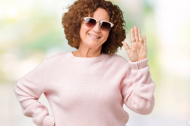 Mulher sênior bonita meio ager vestindo suéter rosa e óculos de sol sobre fundo isolado Renúncia dizendo olá feliz e sorridente, gesto de boas-vindas amigável
 - Foto, Imagem