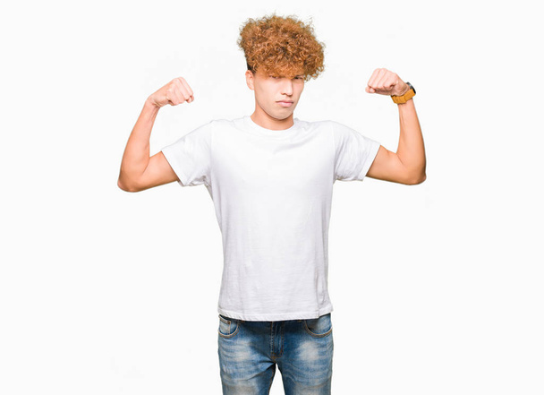 Joven hombre guapo con pelo afro que lleva una camiseta blanca casual que muestra los músculos de los brazos sonriendo orgulloso. Concepto Fitness
. - Foto, Imagen