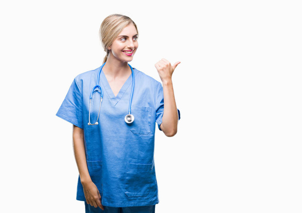 junge schöne blonde Doktor Chirurg Krankenschwester über isolierten Hintergrund lächelnd mit glücklichem Gesicht suchen und zur Seite zeigen mit dem Daumen nach oben. - Foto, Bild