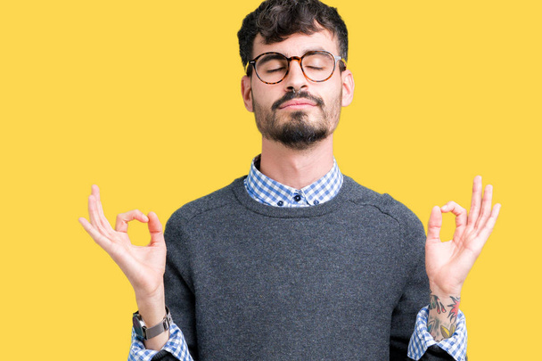 Young handsome smart man wearing glasses over isolated background relax and smiling with eyes closed doing meditation gesture with fingers. Yoga concept. - Photo, Image