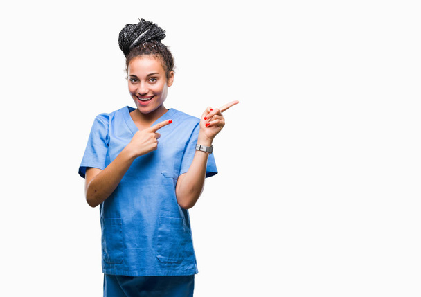 Young braided hair african american girl professional nurse over isolated background smiling and looking at the camera pointing with two hands and fingers to the side. - Photo, Image