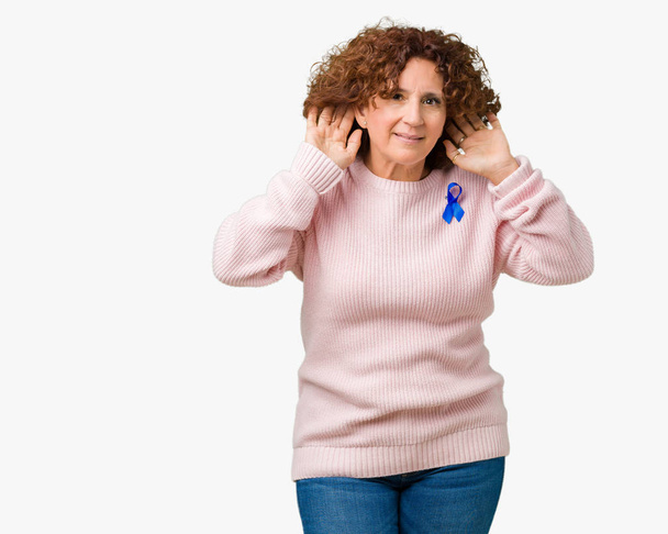 Μεσαίο ager ανώτερων γυναίκα που φοράει ευμετάβλητη μπλε χρώμα κορδέλα ευαισθητοποίησης απομονωμένες φόντο προσπαθεί να ακούω και τα δύο χέρια στο αυτί χειρονομία, περίεργος για κουτσομπολιό. Πρόβλημα ακοής, κουφός - Φωτογραφία, εικόνα