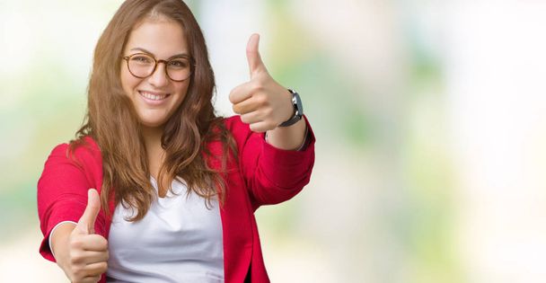 Beautiful plus size young business woman wearing elegant jacket and glasses over isolated background approving doing positive gesture with hand, thumbs up smiling and happy for success. Looking at the camera, winner gesture. - Photo, Image