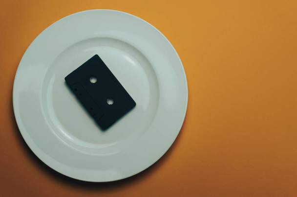 Černá klasická magnetická kazeta v bílém talíři s oranžově zbarvený příbory - Fotografie, Obrázek