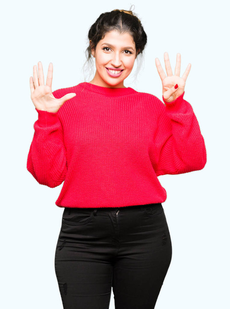 Fiatal szép nő visel piros pulóver és zsemle mutatja, és ujjal felfelé mutató szám kilenc, miközben mosolyogva, magabiztos és boldog. - Fotó, kép