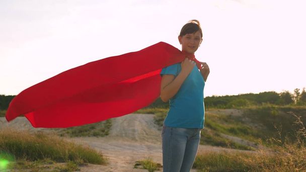 piękna dziewczyna superbohatera stojący na polu w czerwony płaszcz, płaszcz łopocze na wietrze. Zwolnionym tempie. Młoda dziewczyna marzy o zostaniu superbohatera. dziewczyna idzie w wyrażeniu czerwony płaszcz snów - Zdjęcie, obraz