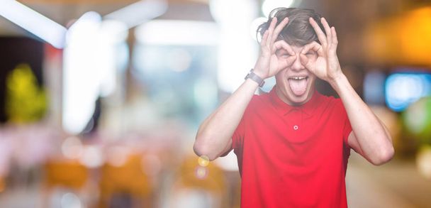 Young handsome man wearing red t-shirt over isolated background doing ok gesture like binoculars sticking tongue out, eyes looking through fingers. Crazy expression. - Photo, Image
