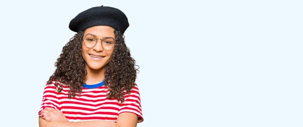 Young beautiful woman with curly hair wearing glasses and fashion beret happy face smiling with crossed arms looking at the camera. Positive person. - Photo, Image