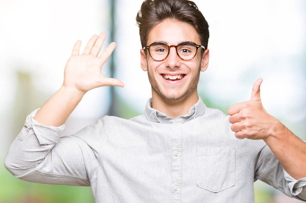 Jovem homem bonito usando óculos sobre fundo isolado mostrando e apontando para cima com os dedos número seis enquanto sorri confiante e feliz
. - Foto, Imagem