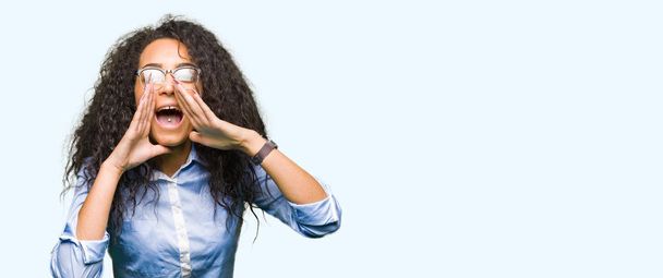 Молодая красивая деловая девушка с вьющимися волосами в очках кричит громко, с руками над ртом
 - Фото, изображение