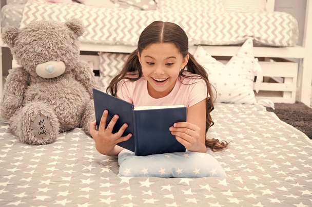 Enjoy favorite moment. Girl child lay bed with teddy bear read book. Kid prepare to go to bed. Girl kid long hair cute pajamas relax and read book to bear toy. Pleasant time in cozy bedroom - Фото, изображение