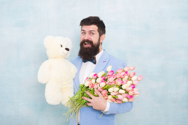 Happy and in love. Gentleman making romantic surprise for her. Gentleman romantic gift. Birthday greetings. Happy birthday my love. Man bearded gentleman suit bow tie hold teddy bear and bouquet - Photo, image