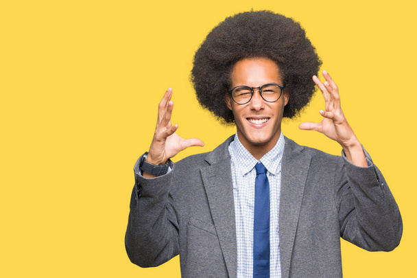 Joven hombre de negocios afroamericano con cabello afro usando gafas Gritando frustrado con rabia, manos tratando de estrangular, gritando loco
 - Foto, imagen