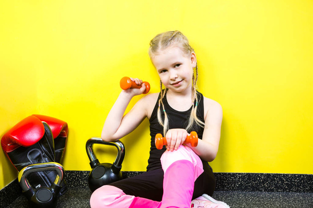 Θεματικά αθλήματα και παιδιά υγείας. Μικρό αστείο παιδί Καυκάσιος κορίτσι με κοτσίδες, κάθεται ανάπαυση διάλειμμα στο πάτωμα στο γυμναστήριο. Αθλητής Αλούλα εξοπλισμοί για γυμναστική bodybuilding φόντο κίτρινο τοίχο - Φωτογραφία, εικόνα