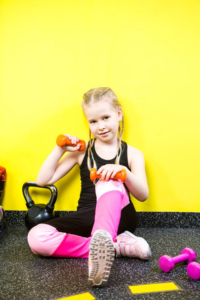 Theme sports and health children. Little funny child Caucasian girl with pigtails, sits resting break on floor in gym. Athlete dumbbell equipments for gymnastics bodybuilding background yellow wall - Photo, Image