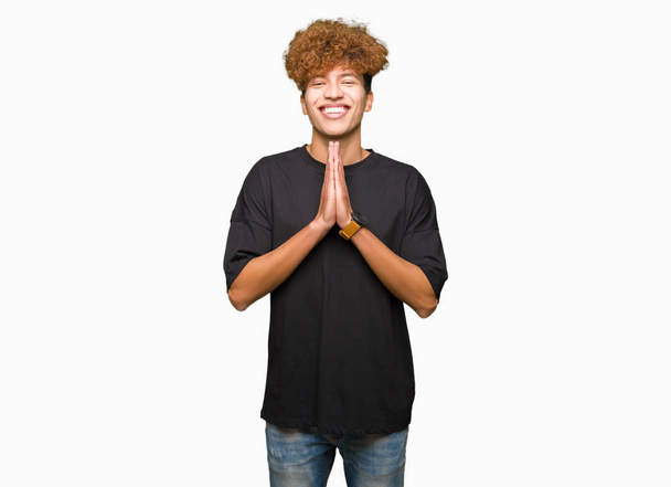Joven hombre guapo con pelo afro vistiendo camiseta negra rezando con las manos juntas pidiendo perdón sonriendo confiado
. - Foto, Imagen