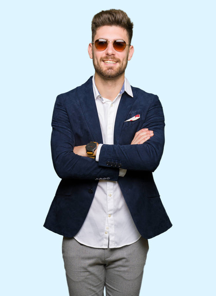 Young handsome elegant man wearing sunglasses and fashion blazer happy face smiling with crossed arms looking at the camera. Positive person. - Foto, afbeelding