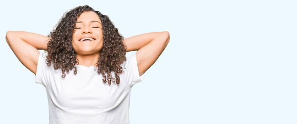 Молодая красивая женщина с вьющимися волосами в белой футболке расслабляется и растягивается с руками и руками за головой и шеей, улыбаясь счастливой
 - Фото, изображение