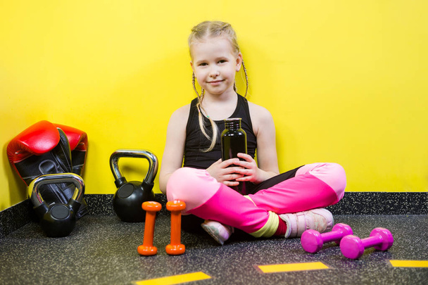 Theme sports and health children. Little Caucasian girl sits resting break floor gym holds hand bottle, drink water thirst. Athlete dumbbell equipments gymnastics bodybuilding background yellow wall - Photo, Image