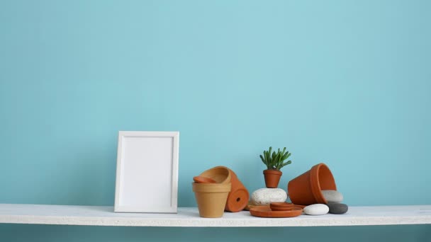 Modern room decoration with picture frame mockup. White shelf against pastel turquoise wall with pottery and succulent plant. Hand putting down potted violet plant. - Footage, Video