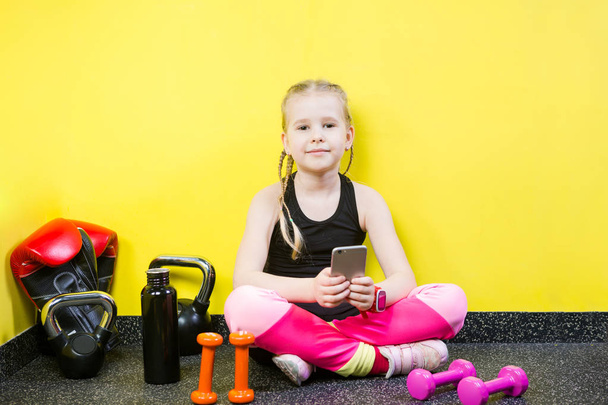 Malá holčička s mobilním telefonem, sedící na podlaze u činek, boxerské rukavice a láhev vody na podlaze. Připraven cvičit v tělocvičně. Sport a zdravý koncept - Fotografie, Obrázek