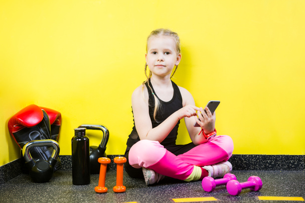 Young child girl with a mobile phone sitting on the floor near the dumbbells, boxing gloves and a bottle of water on the floor. Ready to workout at the gym. Sport and healthy concept - Fotó, kép