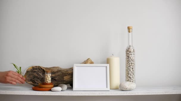 Modern room decoration with picture frame mockup. Shelf against white wall with decorative candle, glass and rocks. Hand putting down potted spider plant. - Footage, Video