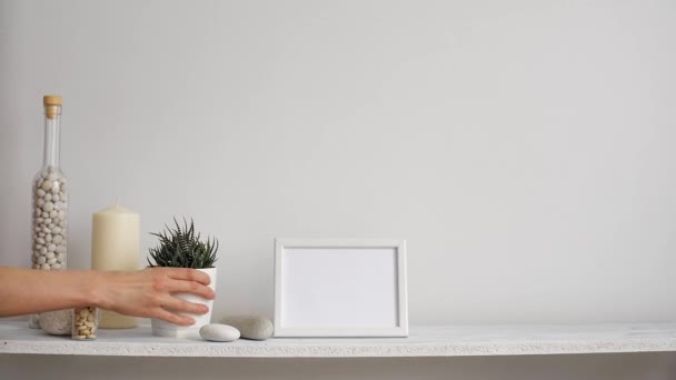 Modern room decoration with picture frame mockup. Shelf against white wall with decorative candle, glass and rocks. Hand putting down potted succulent plant. - Footage, Video