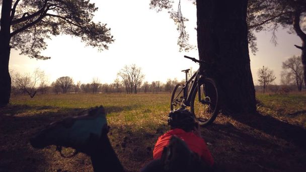 junge kaukasische Athletin Touristenradfahrerin macht Foto am Telefon, fotografiert sein Fahrrad neben Baum auf dem Naturfeld sonnigen Tag stehen. Mädchen mit Helm und Sportbekleidung Ruhepause Mountainbike - Foto, Bild