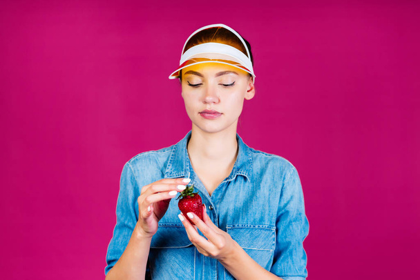 Beautiful girl in a yellow cap eats strawberries on a pink background - Фото, зображення