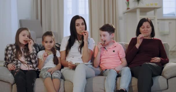 Mãe muito atraente com seus filhos e uma avó comer biscoitos enquanto assiste a uma TV na frente da câmera sentada no sofá em uma espaçosa sala de estar
 - Filmagem, Vídeo