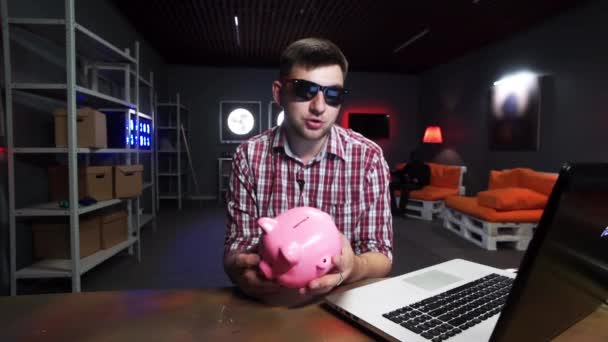 Jonge actieve man met zonnebril houdt plastic varken vast, spreekt en kijkt naar camera - Video