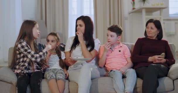 Attractive woman with her three kids and a granny watching movie while eating some biscuits in front of the camera , they have a nice conversation and smiling faces - Felvétel, videó
