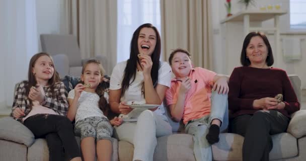 Drie kinderen doorbrengen een leuke tijd met hun moeder en oma kijken naar een komedie op de TV voor de camera ze glimlachen en genieten van de tijd samen zittend op de Bank - Video