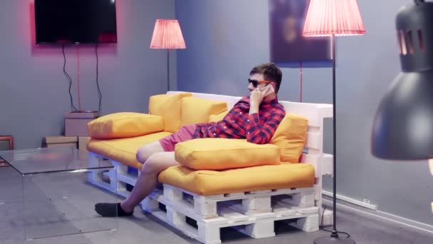 Cute facet z okulary siedzi na pomarańczowej kanapie i mówi na telefon w nowoczesnym pokoju - Materiał filmowy, wideo