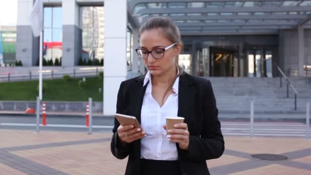 スマートフォンを持つ若いビジネス女性はオフィスに対して街の通りにコーヒーを飲みます。安定したショット - 映像、動画