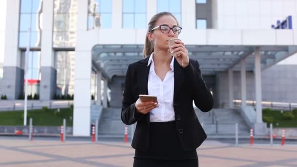 Mujer de negocios bonita con teléfono inteligente beber café en la calle de la ciudad contra la oficina. Disparo constante
 - Metraje, vídeo