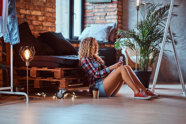 Curly blond woman is sitting next to the pallet sofa while looking to her smartphone.  - Foto, immagini