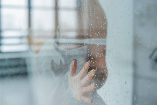 Young woman, posing behind transparent glass covered by water drops. melancholy and sad female portrait. Blonde in a white t-shirt touches the glass with drops in the shower room - Foto, immagini