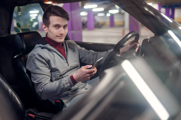 Photo of side of smiling male driver with phone in hands sitting in car on underground insurance - Valokuva, kuva