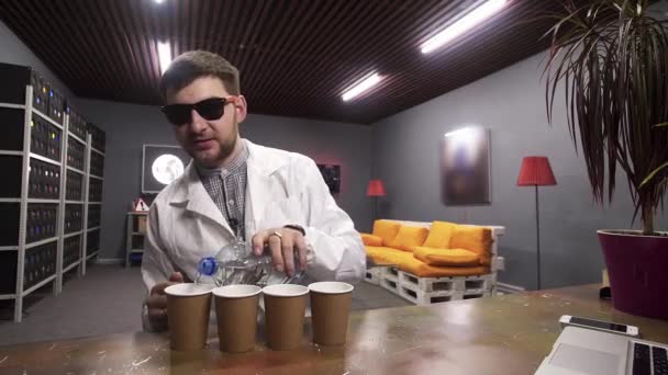 Cute guy in white lab coat pours water from plastic bottle to four paper cups - Footage, Video